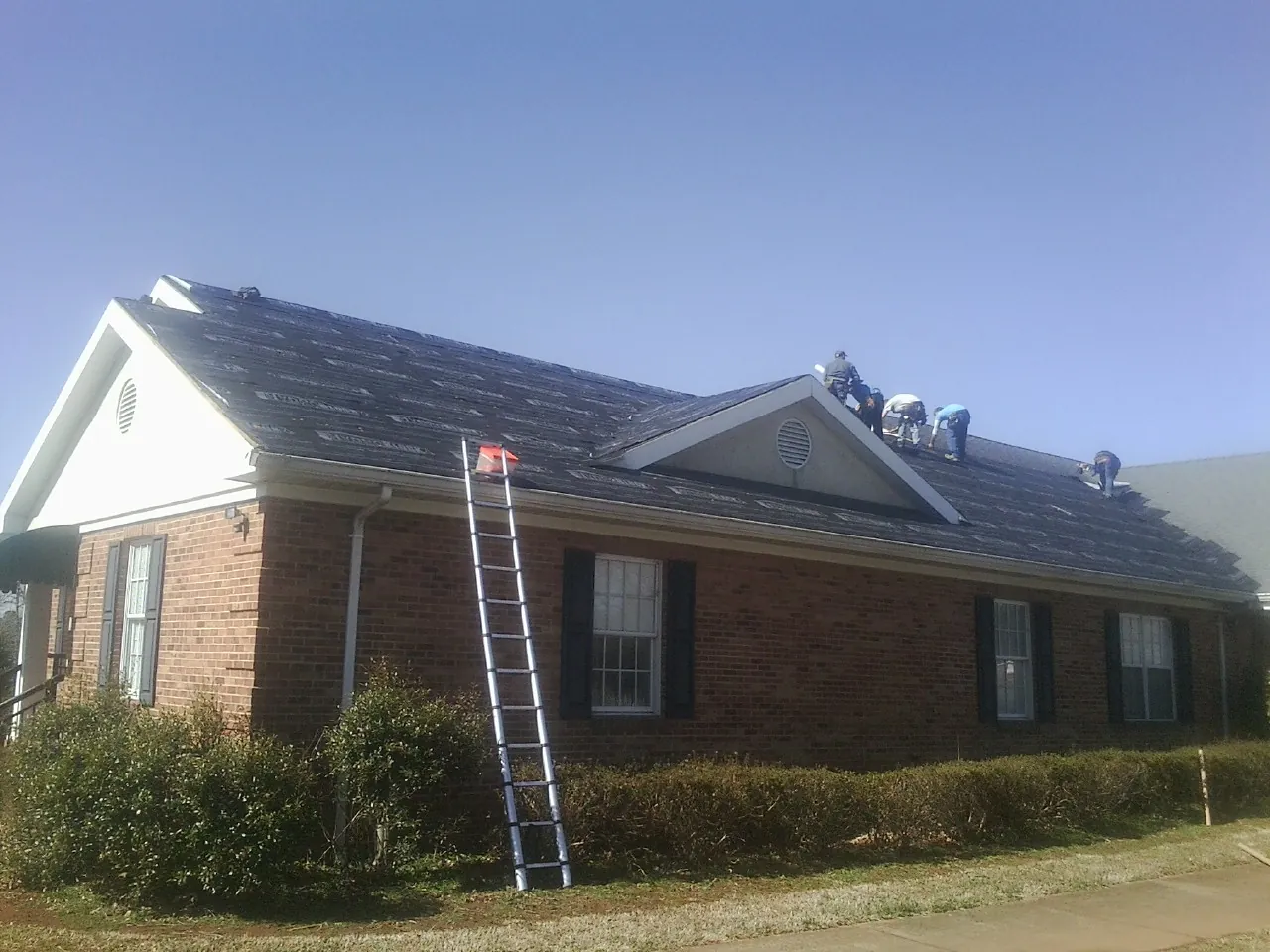 Crew tearing shingles off a brick ranch roof with a ladder set — Metal Roof Repair work by our crew in Buford
