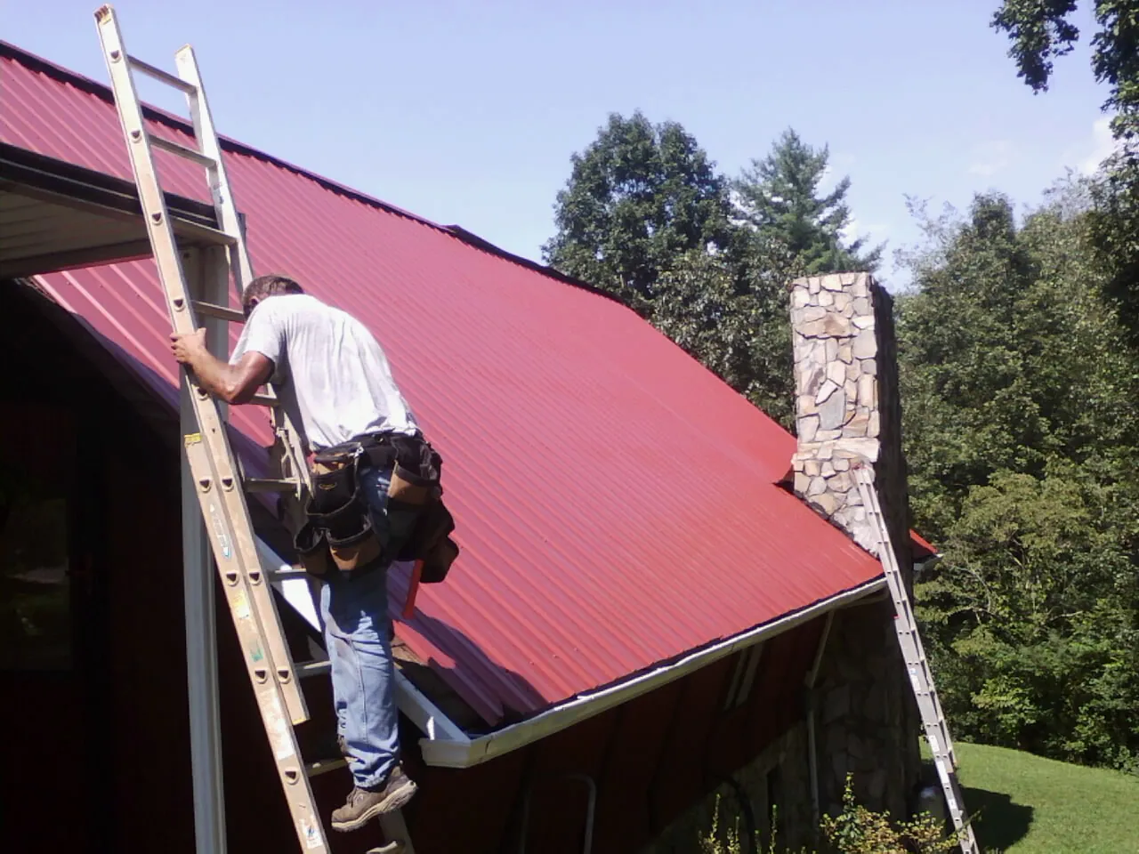 Roofer on a ladder beside a red metal roof and stone chimney — Soffit & Fascia Repair by Roofing Brothers in Buford
